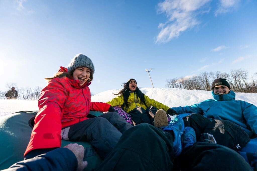 A group of friends laughing and holding hands as they go down hill on a group of snow tubes